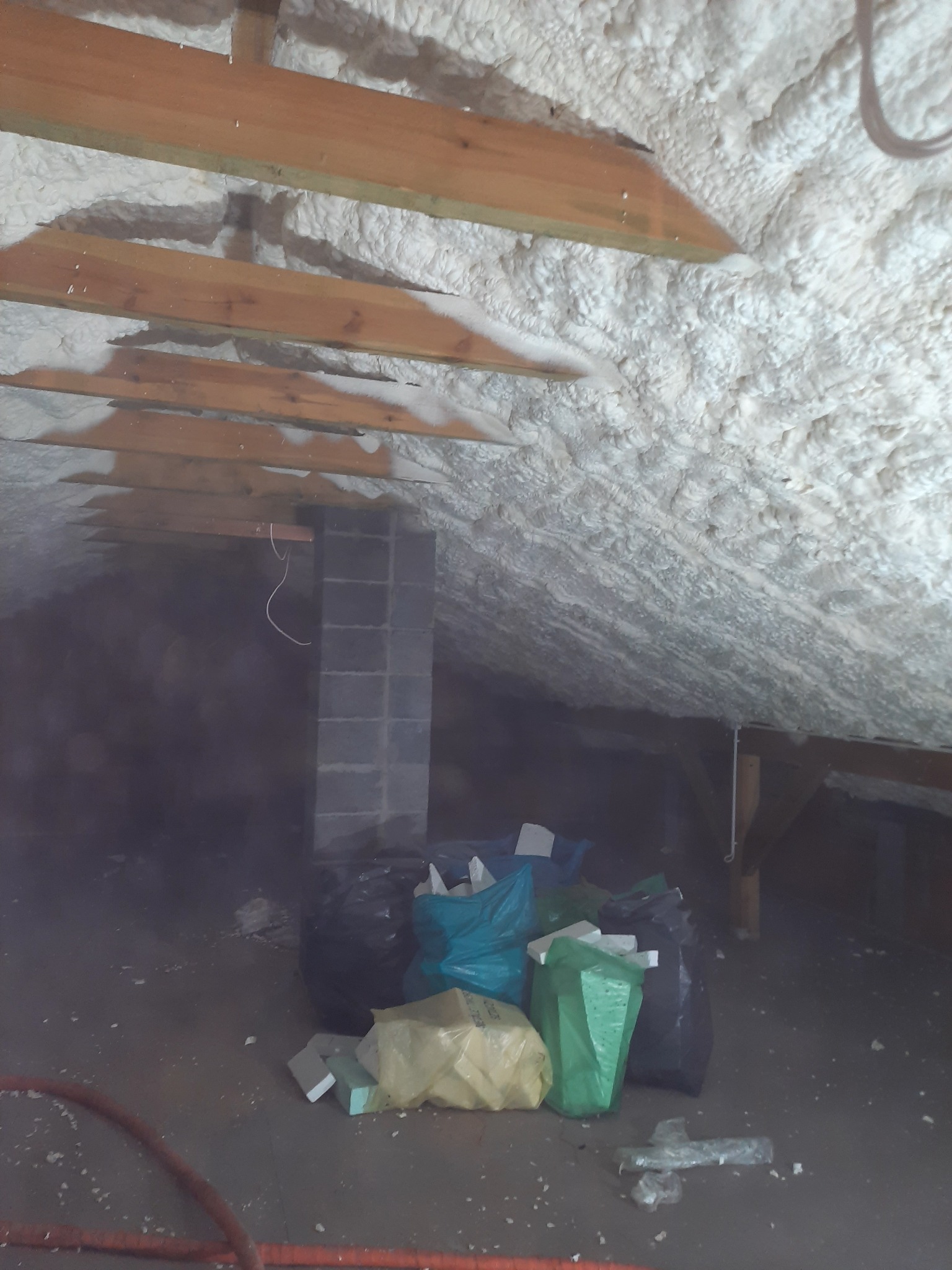 Poddasze po ociepleniu pianką: widoczne drewniane belki stropowe, szara ściana kominowa i worki z odpadami budowlanymi na podłodze.