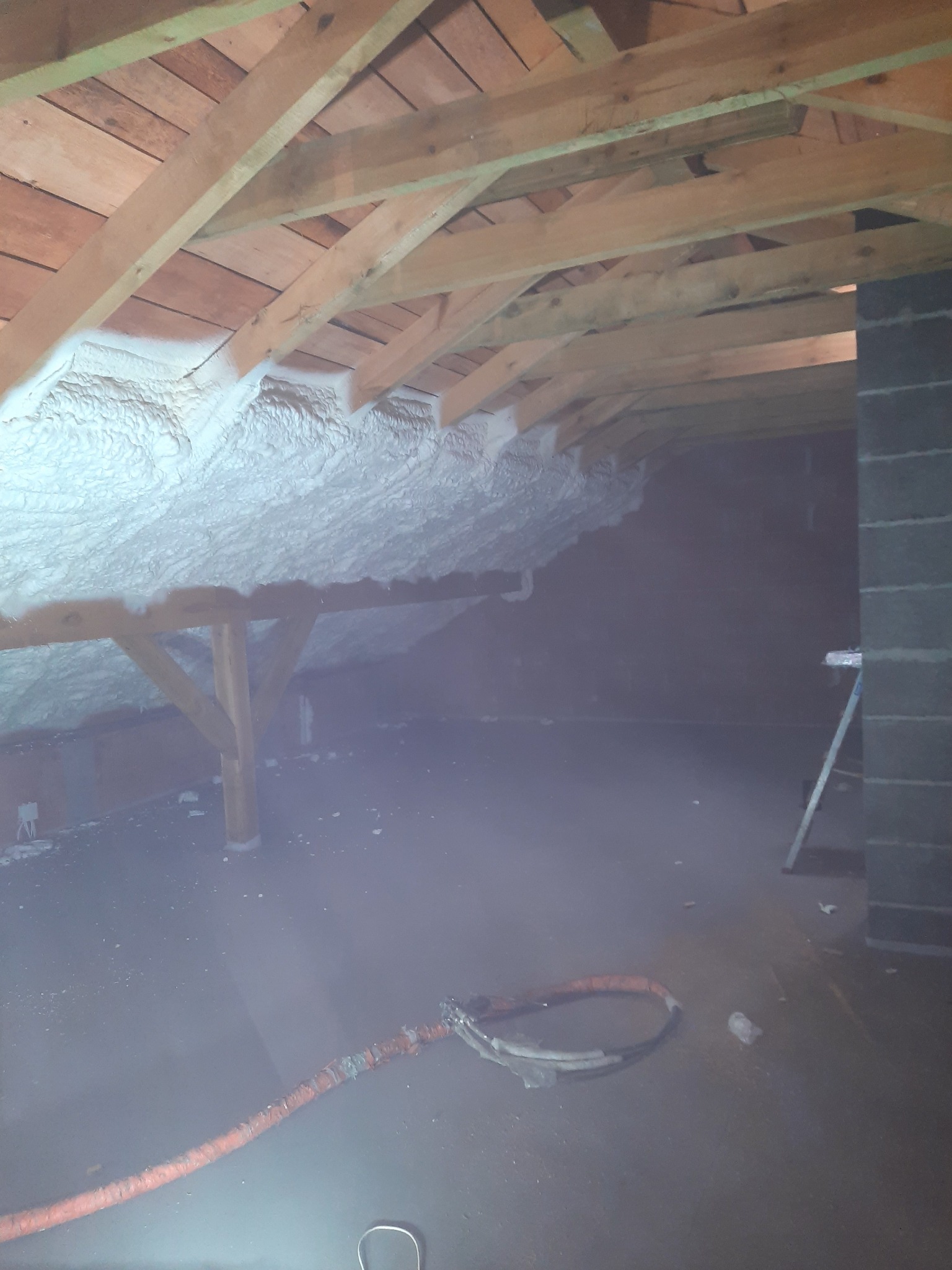 Poddasze z drewnianą konstrukcją dachu, częściowo pokryte białą pianką PUR, widoczny wąż do aplikacji pianki na podłodze i drabina przy ścianie z pustaków.