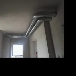 Dwie równoległe rury spiro, zabezpieczone termicznie, zamontowane pod sufitem w surowym wnętrzu budynku w trakcie wykończenia.