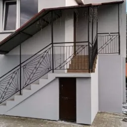 Dwukondygnacyjny budynek z nową, jasnoszarą elewacją, schodami zewnętrznymi z czarną, ozdobną balustradą i zadaszeniem, widok z poziomu podwórka.