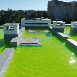 Zielona, płaska powierzchnia dachu z elementami wentylacyjnymi i obróbkami blacharskimi w słoneczny dzień.