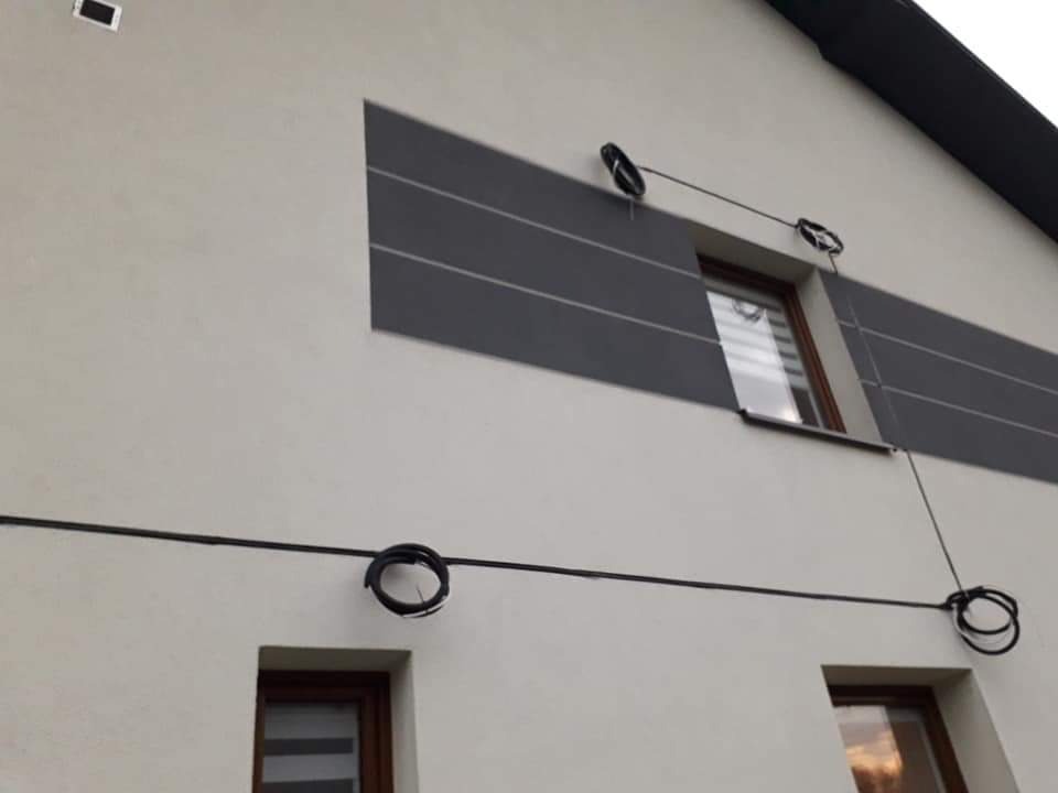 Elewacja domu z widocznymi kablami elektrycznymi prowadzonymi na zewnątrz, przymocowanymi do ściany za pomocą uchwytów, z oknami w tle.