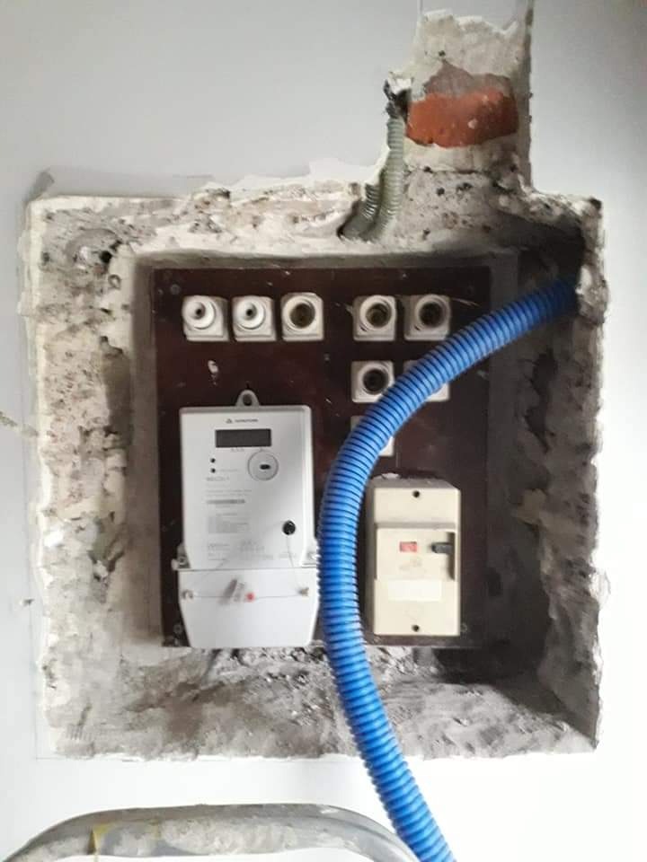 Wnęka w ścianie z zamontowaną tablicą bezpiecznikową, licznikiem energii elektrycznej i niebieską rurką karbowaną, widoczne ślady po kuciu i niedokończone wykończenie ściany.