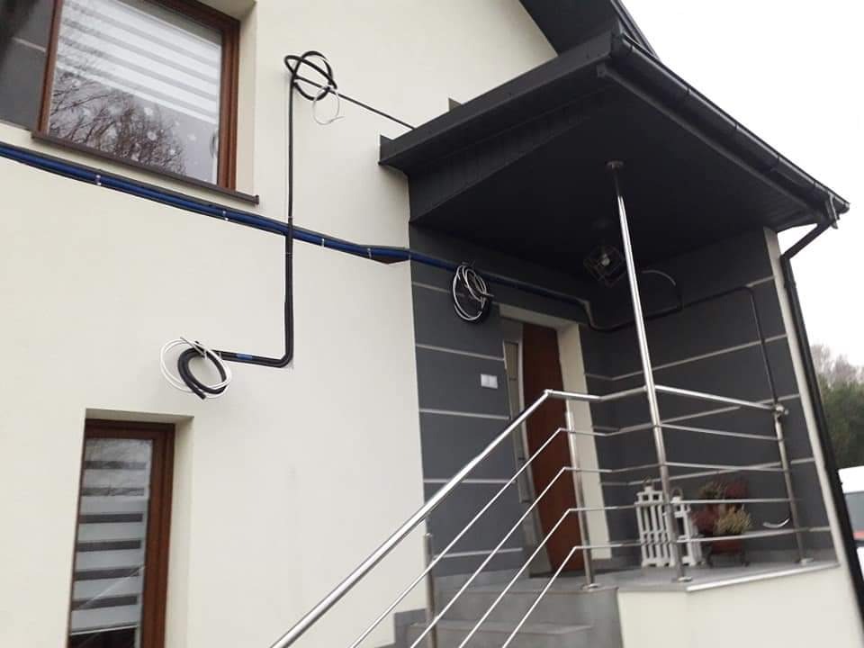 Zewnętrzna instalacja elektryczna na elewacji domu z widocznymi przewodami w peszlach mocowanych do ściany, obok okna i nad wejściem.