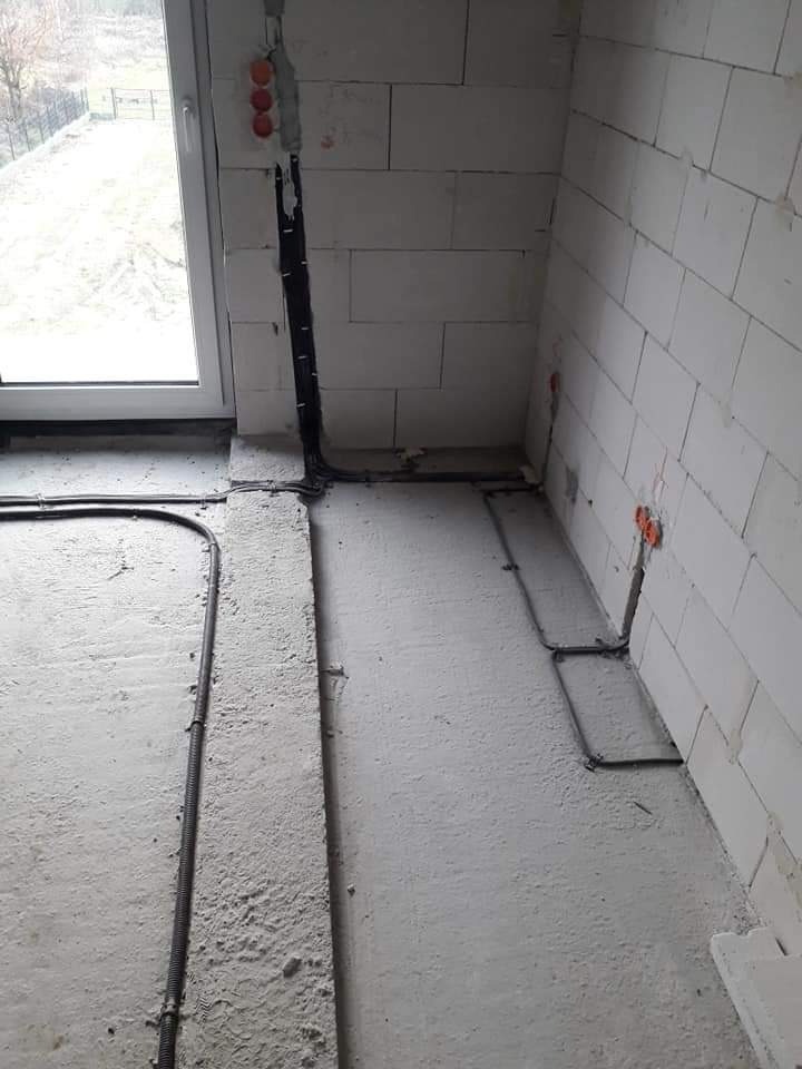 Surowy stan: Widok na wnętrze w trakcie prac, widoczne przewody elektryczne prowadzone po podłodze i ścianach z białego betonu komórkowego, puszki elektryczne w ścianie, okno w tle.