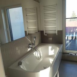 S Burakovskyi - Nowoczesna, biała wanna z hydromasażem, zamontowana pod oknem z widokiem na taras, z dwoma grzejnikami drabinkowymi na ścianie wyłożonej mozaiką w odcieniach szarości.