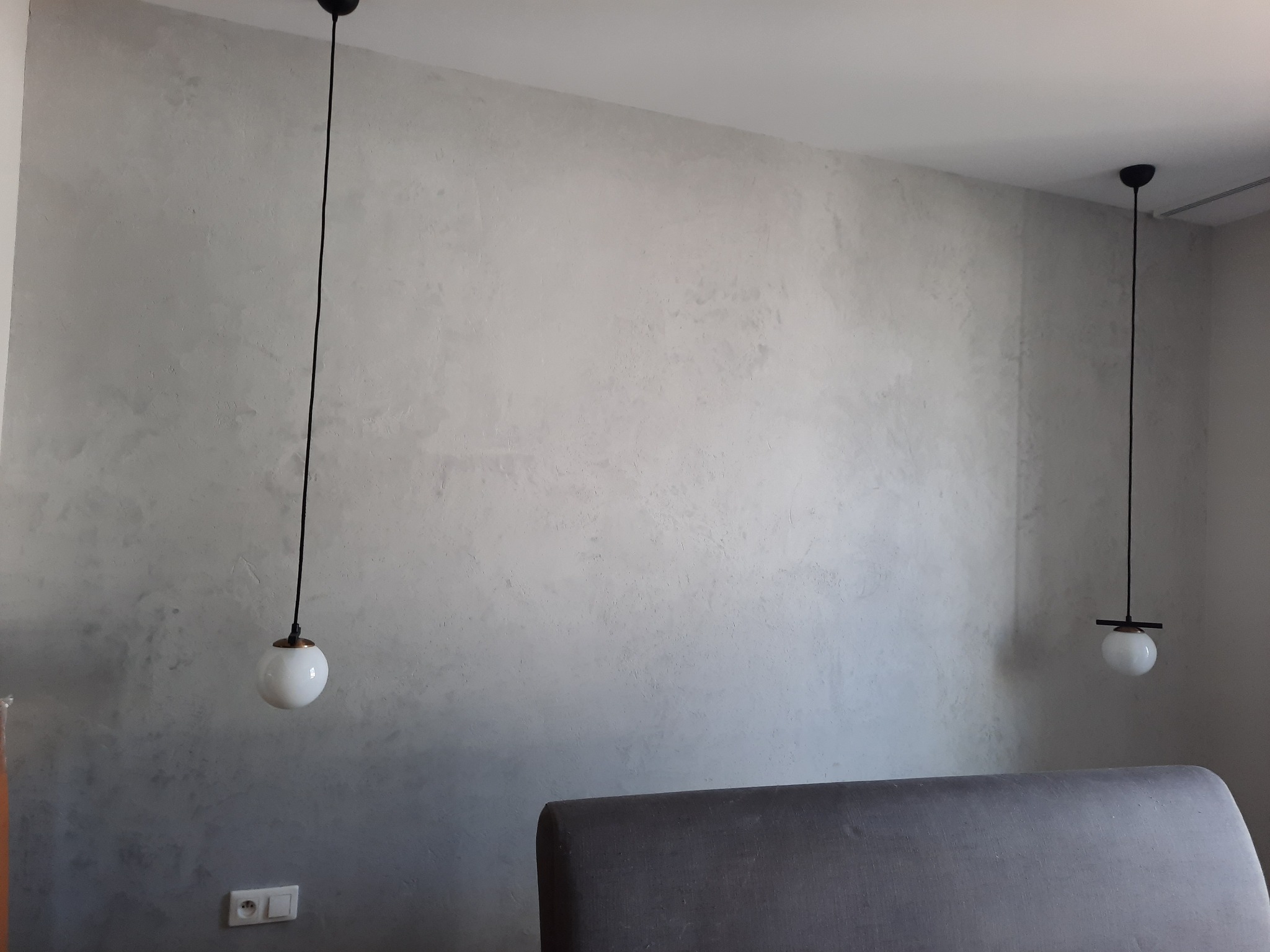 Ściana wykończona szarą gładzią dekoracyjną z dwoma minimalistycznymi lampami wiszącymi z białymi kulistymi kloszami, widoczny fragment szarego zagłówka łóżka.