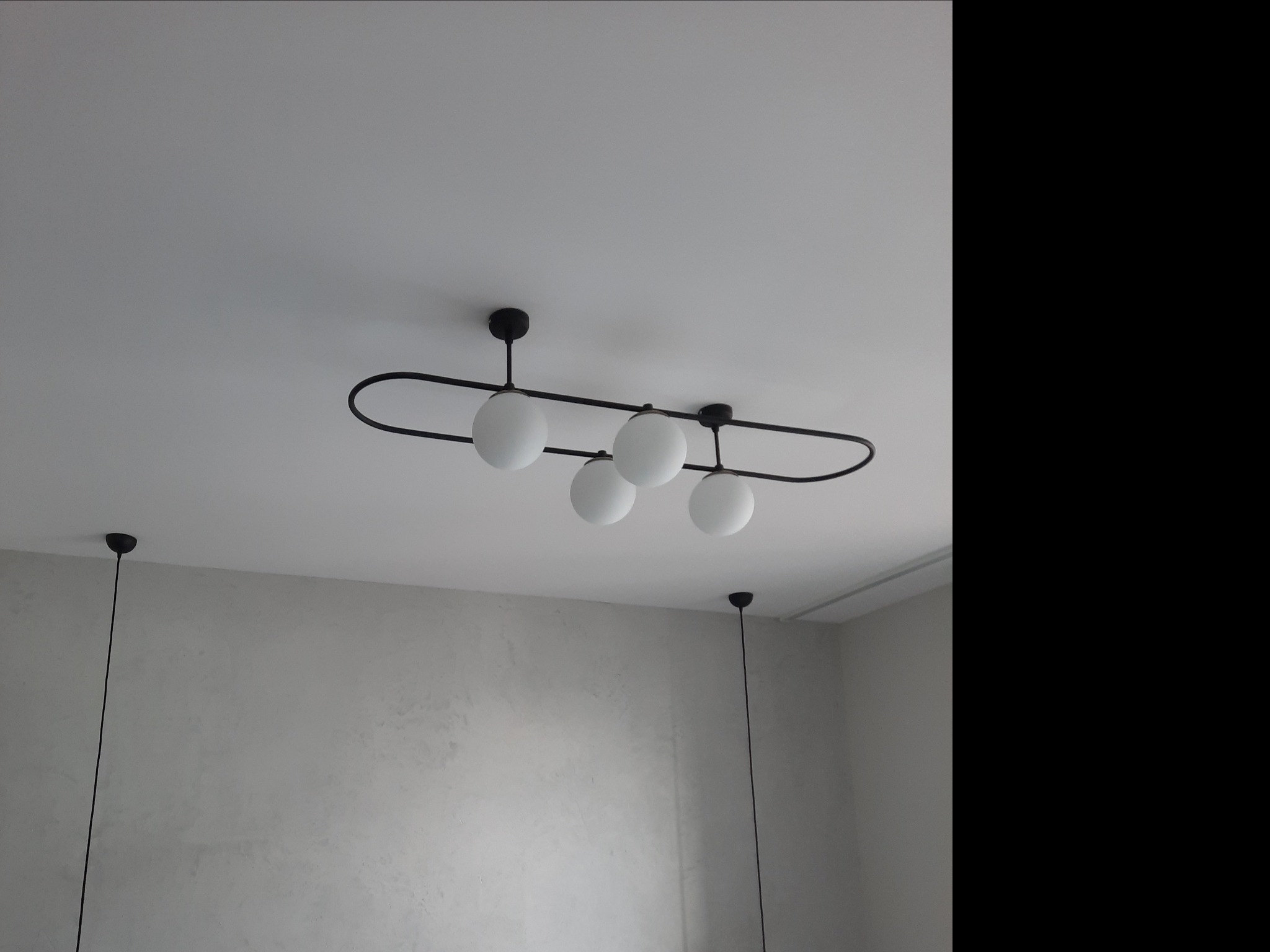 Nowoczesny, czarny żyrandol z czterema białymi, okrągłymi kloszami zawieszony na białym suficie w pokoju z szarą, teksturowaną ścianą. W tle widoczna lampa wisząca na pojedynczym kablu.