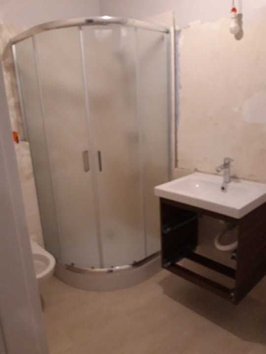 Łazienka w trakcie remontu: kabina prysznicowa z matowego szkła, częściowo zamontowana szafka pod umywalkę z widocznymi rurami i surowa ściana z widocznymi śladami po instalacji elektrycznej.