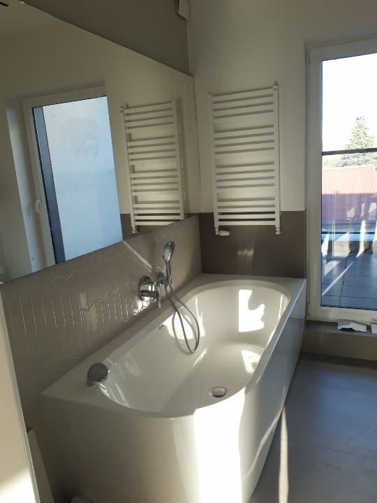 Nowoczesna, biała wanna z hydromasażem, zamontowana pod oknem z widokiem na taras, z dwoma grzejnikami drabinkowymi na ścianie wyłożonej mozaiką w odcieniach szarości.