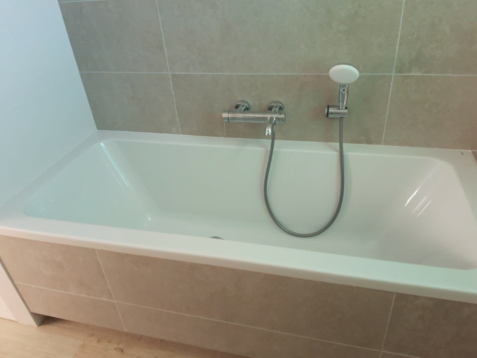 Biała wanna obudowana płytkami imitującymi kamień, z zamontowaną baterią i słuchawką prysznicową na tle ściany wyłożonej płytkami w podobnym odcieniu.