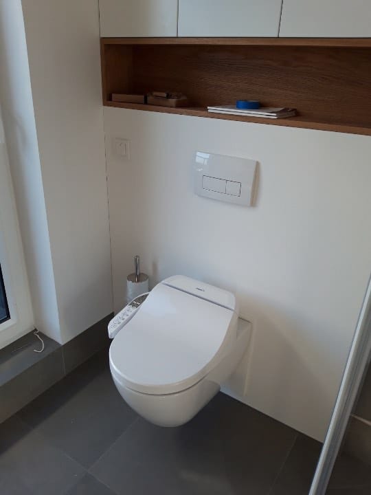 Biała toaleta myjąca z panelem sterowania zamontowana na ścianie w nowoczesnej łazience z szarą podłogą i drewnianą półką nad spłuczką.