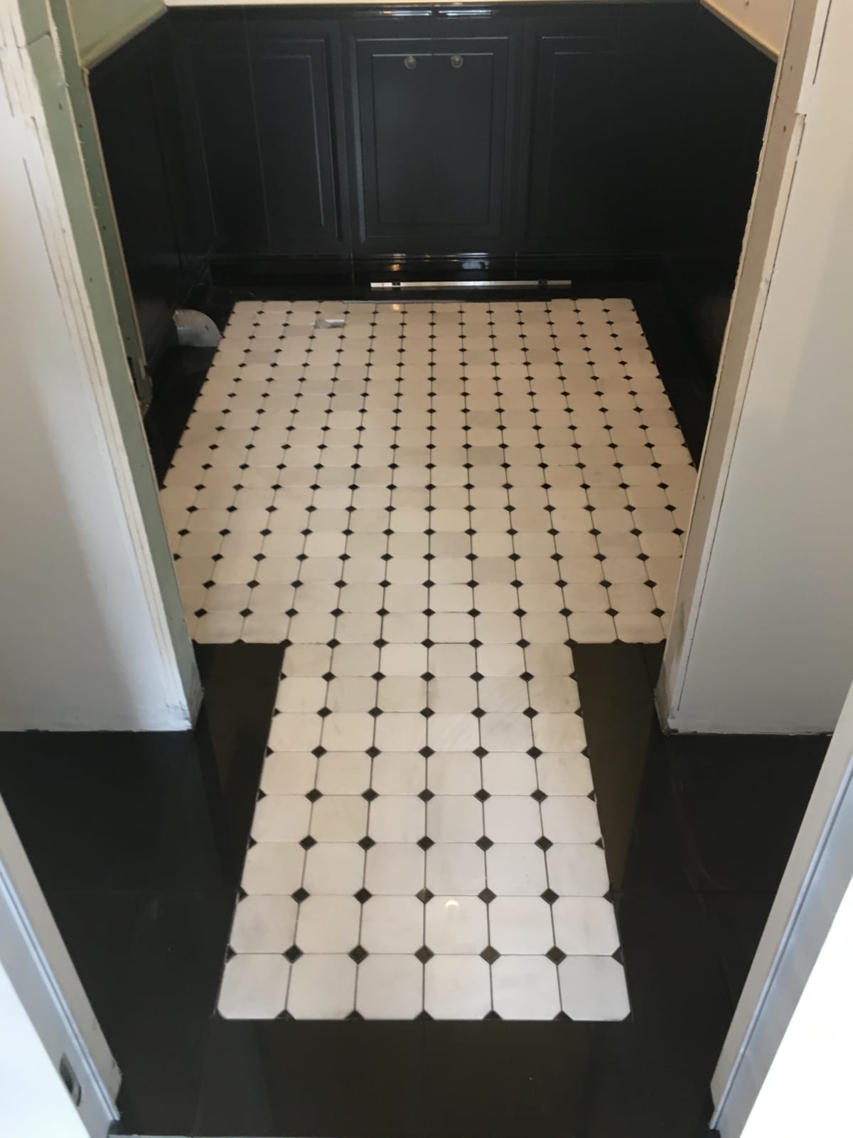 Ułożona podłoga z białych kwadratowych płytek z czarnymi akcentami w wykończonym wnętrzu z czarnymi szafkami w tle.