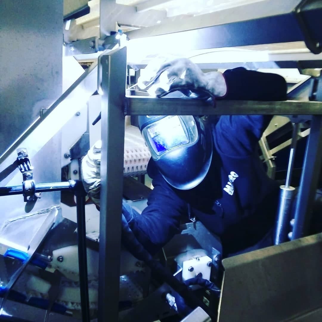 Pracownik w masce spawalniczej, spawający metalową konstrukcję w warsztacie, widoczne iskry i elementy maszyny.