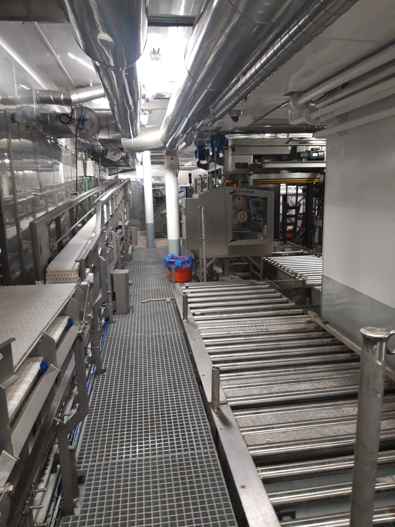 Wnętrze hali produkcyjnej z systemem transportu rolkowego i kratami podłogowymi, widoczne rury wentylacyjne pod sufitem i elementy maszyn.