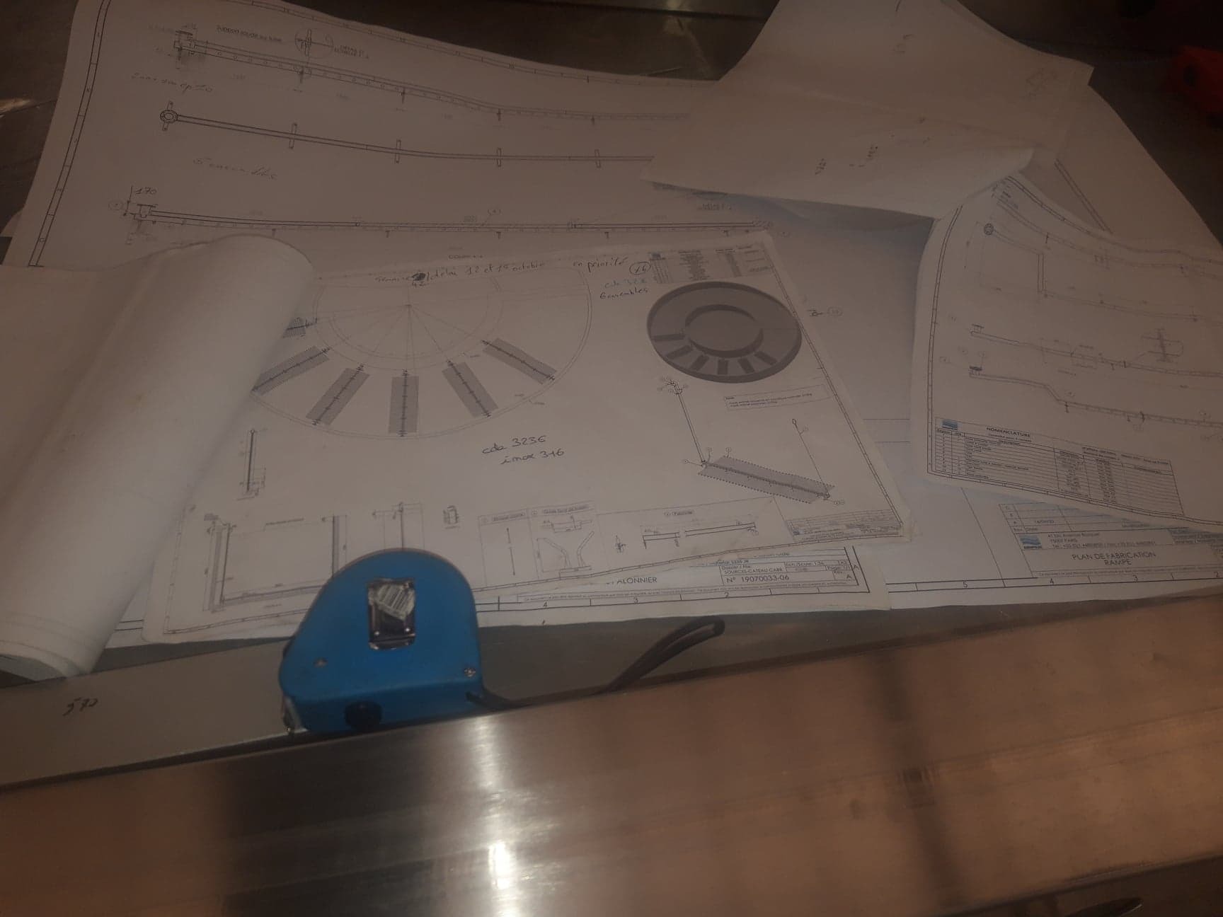 Rozłożone plany techniczne na metalowym stole, z widoczną miarką zwiniętą w niebieskiej obudowie, prezentujące schematy i wymiary konstrukcji metalowych.