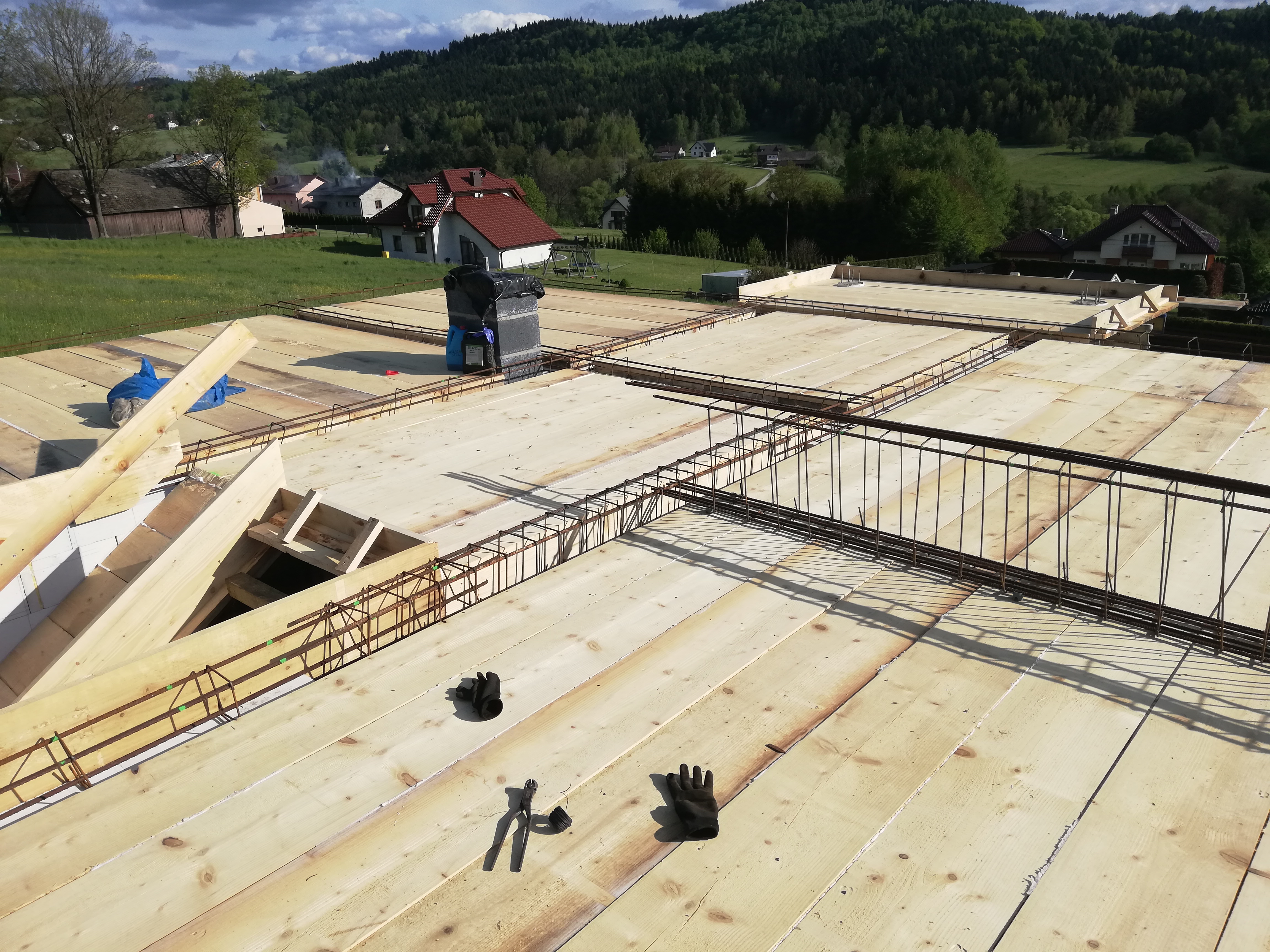 Wiązanie zbrojenia na drewnianym szalunku stropu, widoczne narzędzia i rękawice robocze, w tle krajobraz wiejski z domami i lasem.