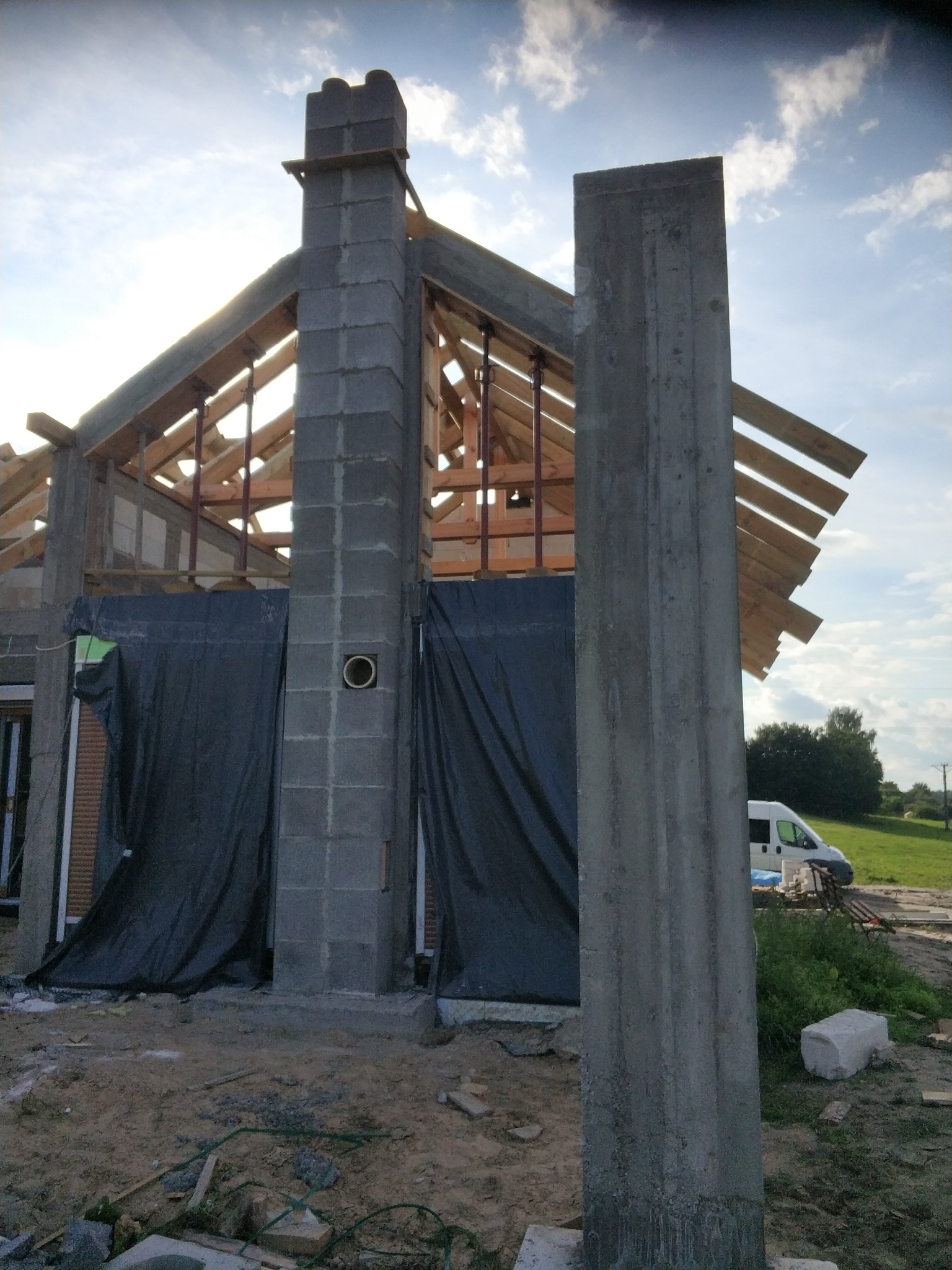 Budowa domu w toku: wzniesiony komin z pustaków szalunkowych z otworem wentylacyjnym, widoczna konstrukcja dachu i betonowy słup na tle nieba.
