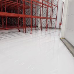 TD FLOOR TOMASZ OCHMAŃSKI - Błyszcząca, biała powierzchnia w magazynie z czerwonymi regałami. Widoczne odbicia w posadzce. Minimalistyczny, industrialny wygląd.