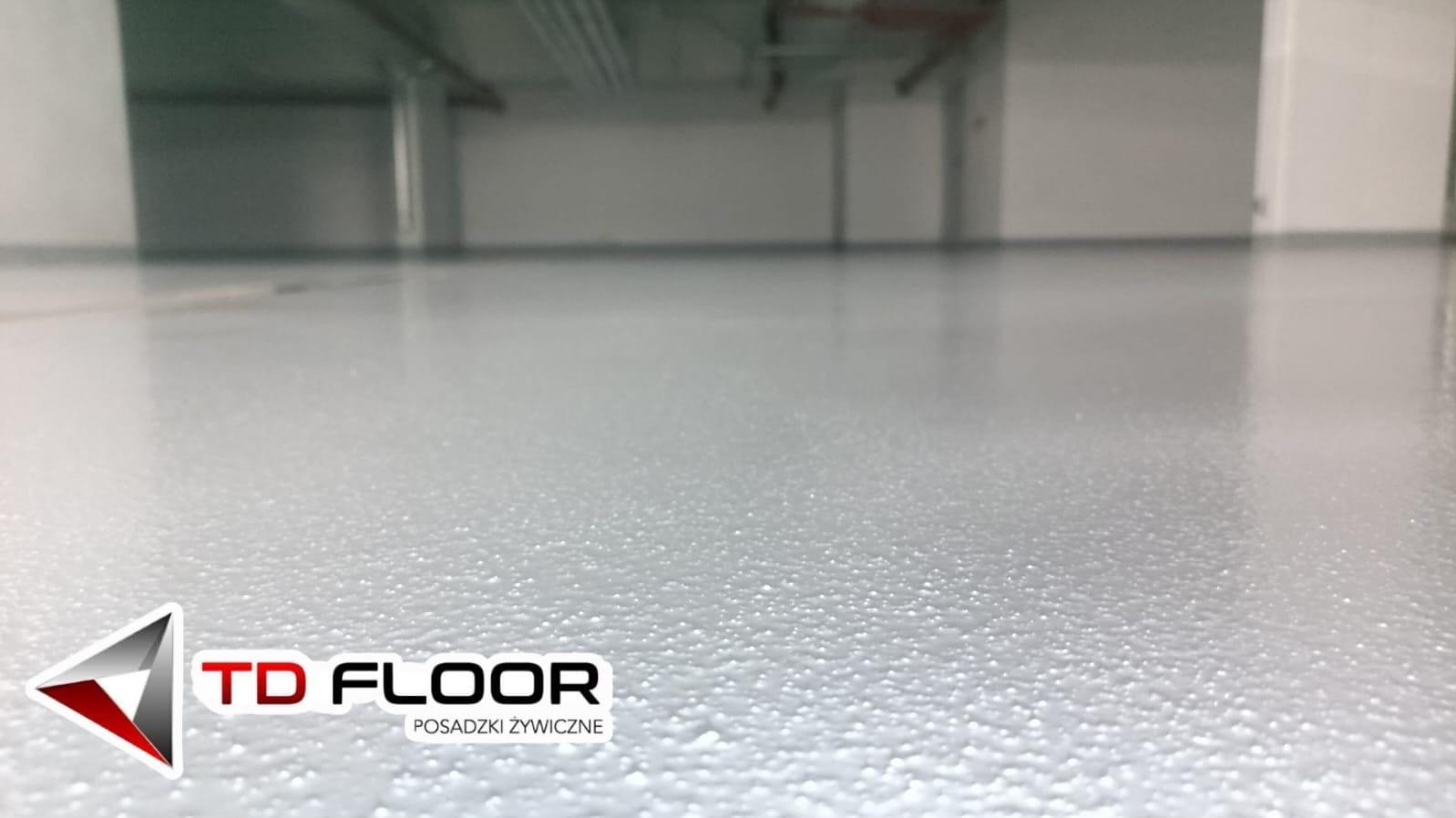 Jasnoszara, gładka powierzchnia w hali, wykonana z żywicy, z widocznym logo firmy TD Floor w lewym dolnym rogu. Widoczne rury pod sufitem.
