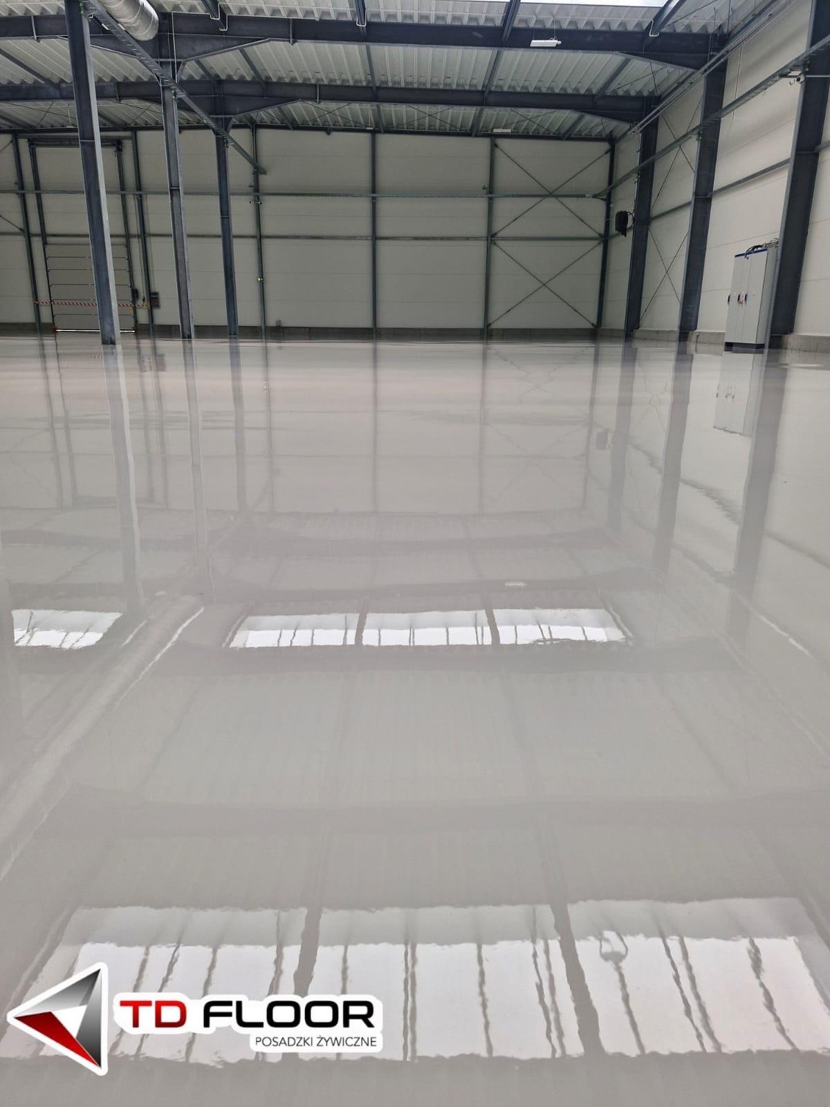 Błyszcząca, jasnoszara posadzka w hali przemysłowej z widoczną konstrukcją stalową dachu i ścian. Gładka powierzchnia odbija światło, tworząc lustrzany efekt. Widoczny logotyp firmy.