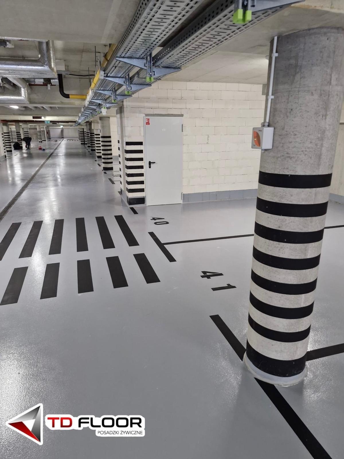Szara posadzka żywiczna w garażu podziemnym z czarnymi oznaczeniami miejsc parkingowych i numerami, kolumny w czarno-białe pasy, instalacje pod sufitem.