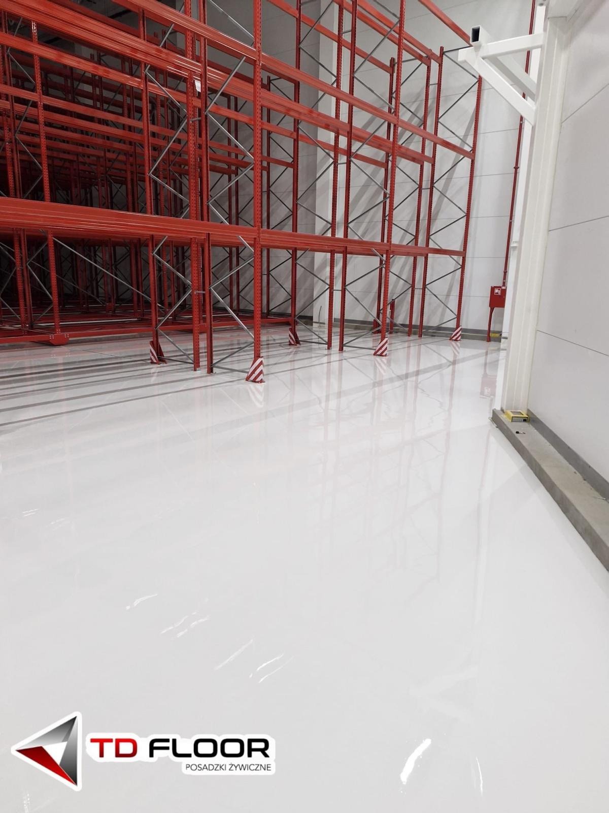 Błyszcząca, biała powierzchnia w magazynie z czerwonymi regałami. Widoczne odbicia w posadzce. Minimalistyczny, industrialny wygląd.