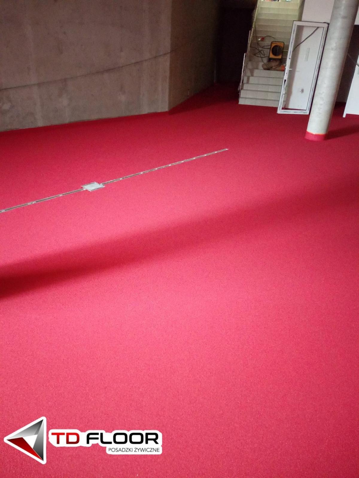 Czerwona, matowa posadzka w hali, z widocznym metalowym profilem i fragmentem schodów w tle. Gładka powierzchnia, jednolite wykończenie, surowe ściany.