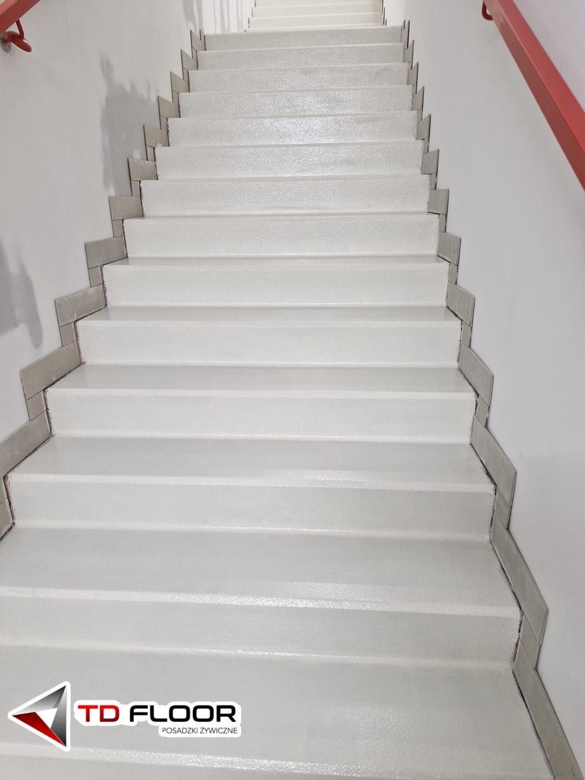 Białe schody z żywicy, wykończone szarymi płytkami na krawędziach, prowadzące w górę. Czerwona poręcz po prawej stronie. Widoczny logotyp firmy w dolnej części zdjęcia.