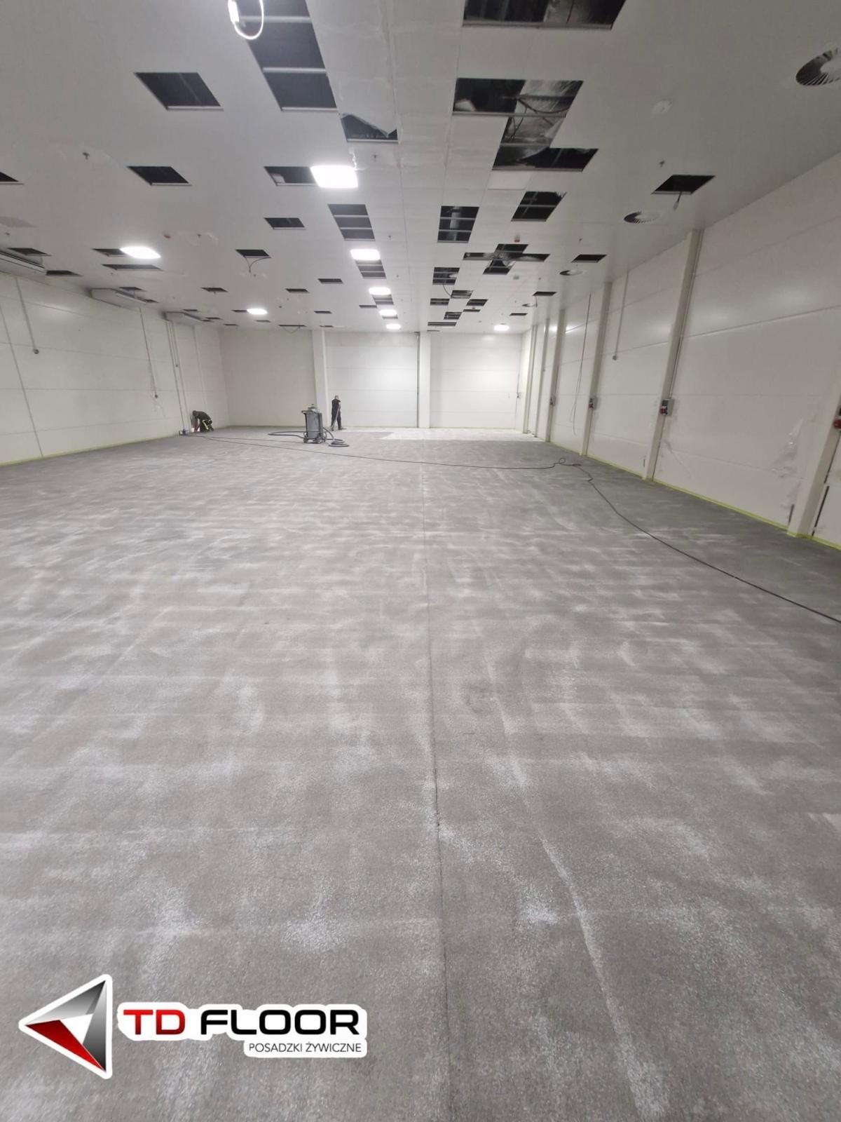 Szara, przemysłowa powierzchnia w trakcie przygotowania pod wylanie. Widoczni dwaj pracownicy i sprzęt. Sufit z otworami na oświetlenie.