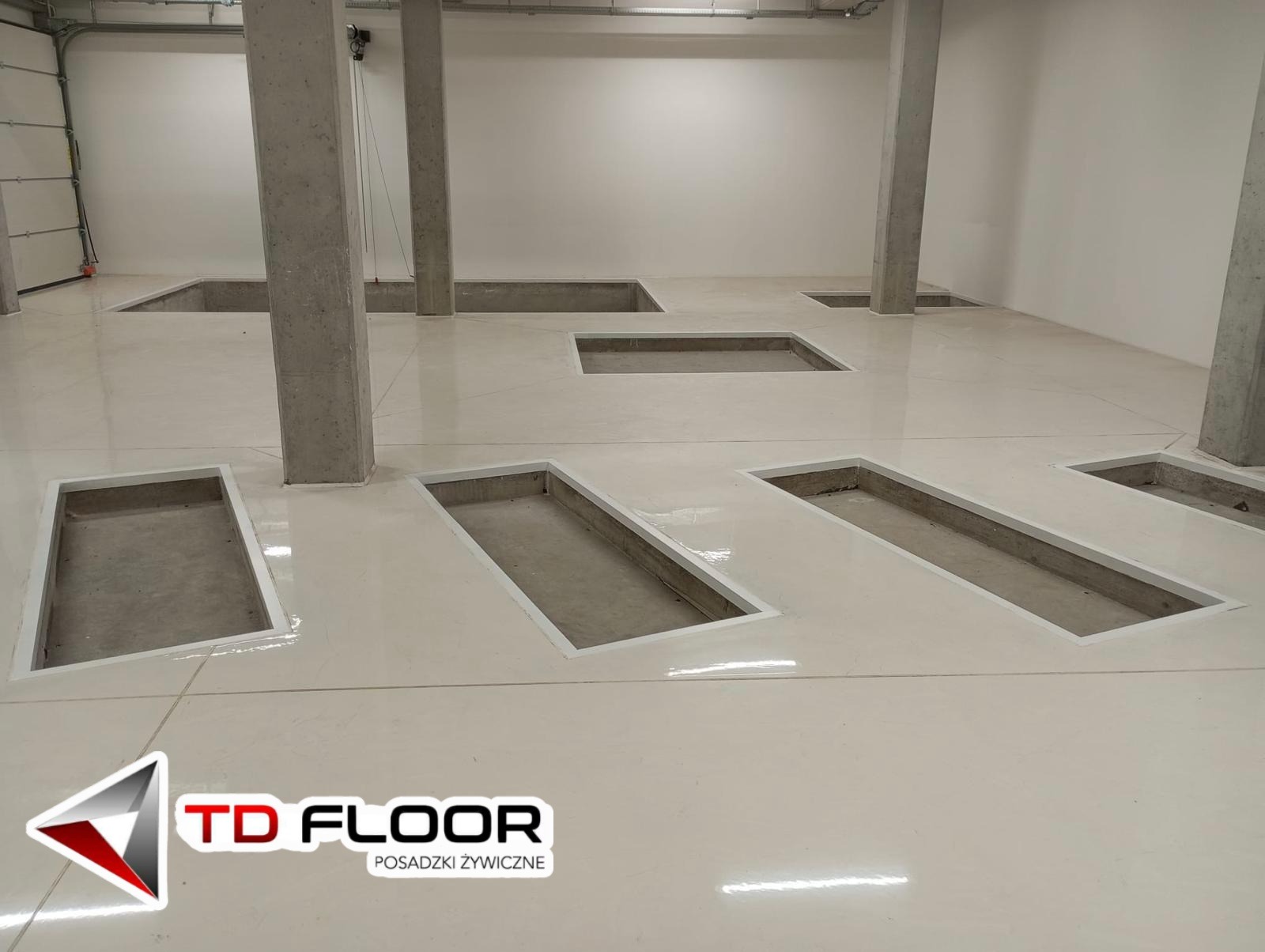 Jasna, błyszcząca posadzka w hali z kanałami rewizyjnymi, wykończona białą listwą. Widoczne betonowe filary i brama garażowa w tle.