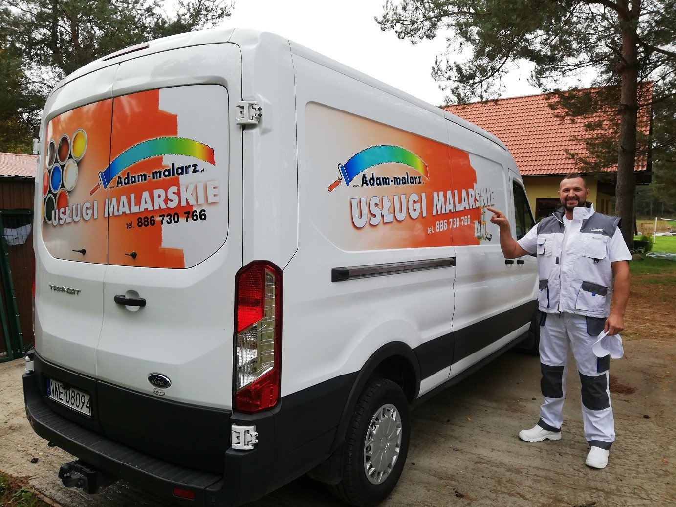 Biały samochód dostawczy z reklamą usług malarskich 'Adam-malarz' i numerem telefonu, obok którego stoi mężczyzna w ubraniu roboczym wskazujący na reklamę.