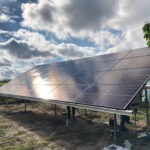 Panel słoneczny ustawiony na metalowej konstrukcji na otwartym terenie, odbijający chmurne niebo.