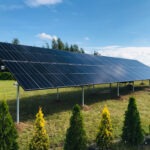 Instalacja paneli słonecznych na konstrukcji wsporczej, widoczna na tle zielonego trawnika i drzew. Panele ustawione pod kątem, odbijają błękitne niebo.