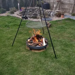 Metalowe palenisko ogrodowe na trójnogu z rusztem, płonące drewno w palenisku, w tle trawnik, kamienny ogródek i ogrodzenie.