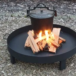 Czarny, metalowy palenisko ogrodowe z palącym się drewnem i wiszącym nad nim kociołkiem, ustawione na żwirowym podłożu.