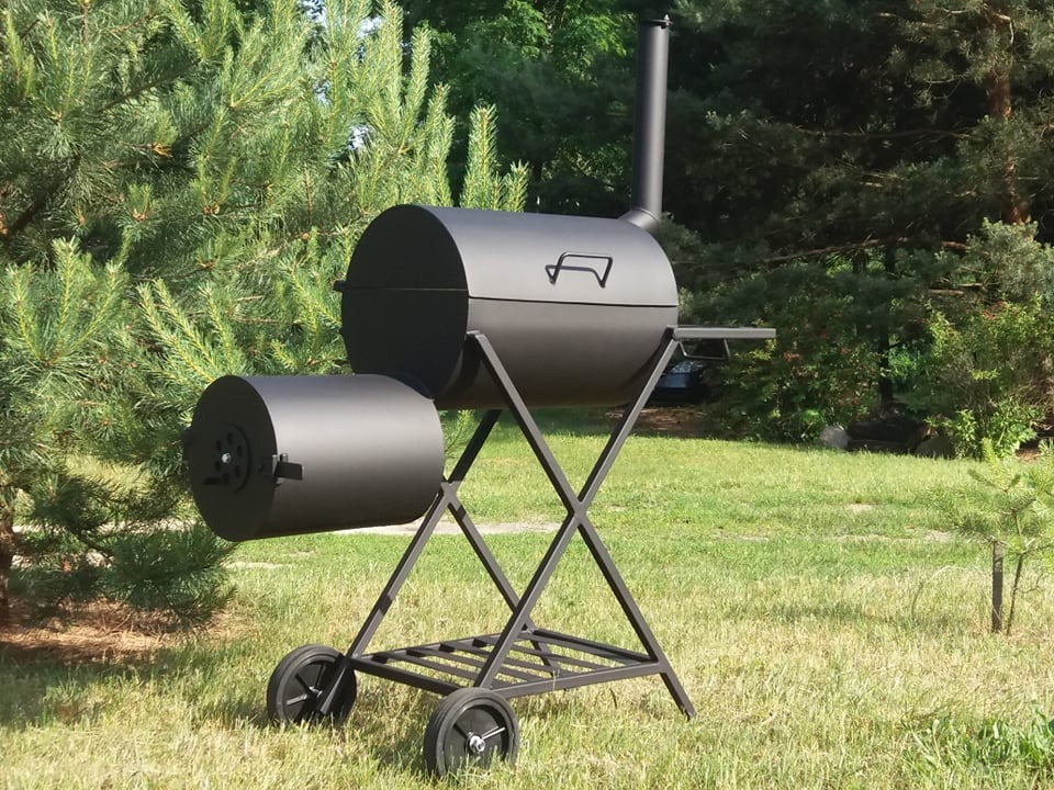 Czarny, metalowy grill wędzarniczy z bocznym paleniskiem, umieszczony na trawniku przed drzewami.