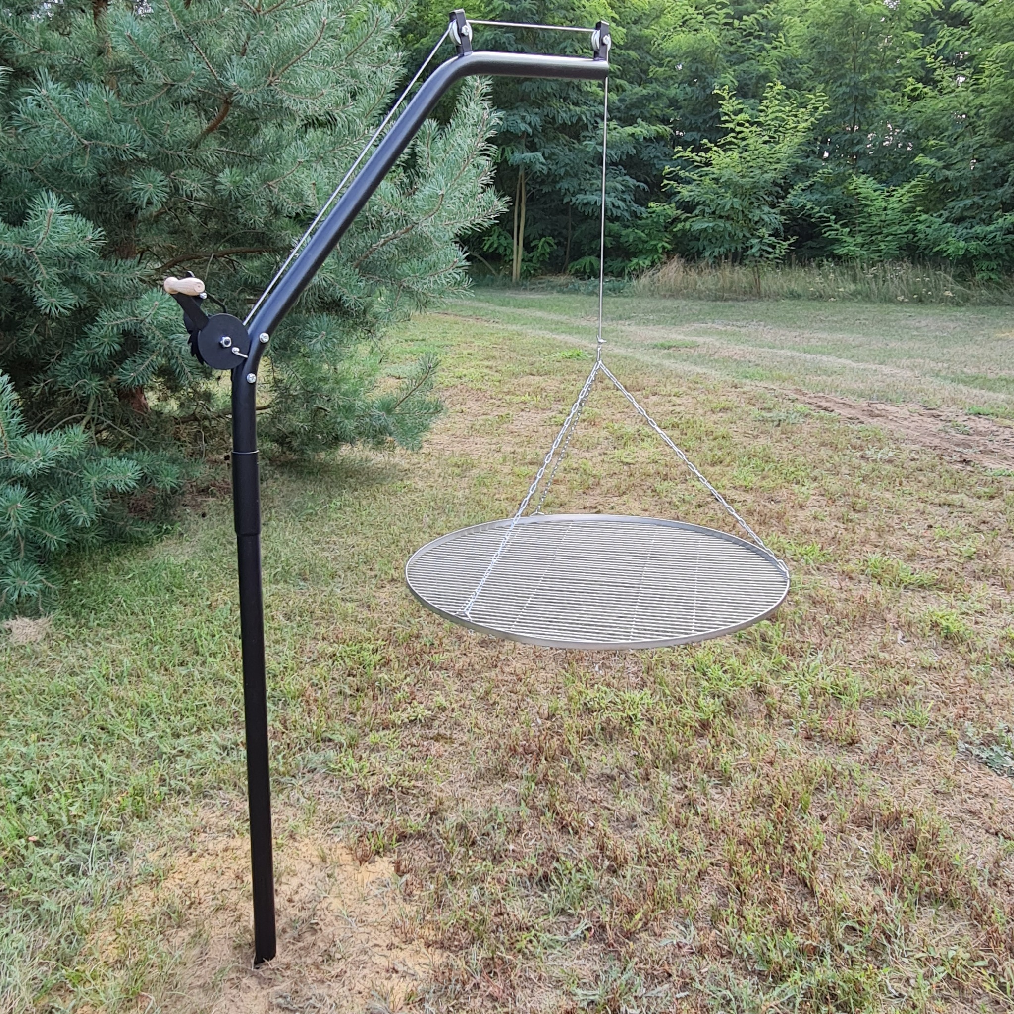Metalowy grill wiszący na regulowanym, czarnym stelażu, umieszczony na trawiastym terenie z drzewami w tle.