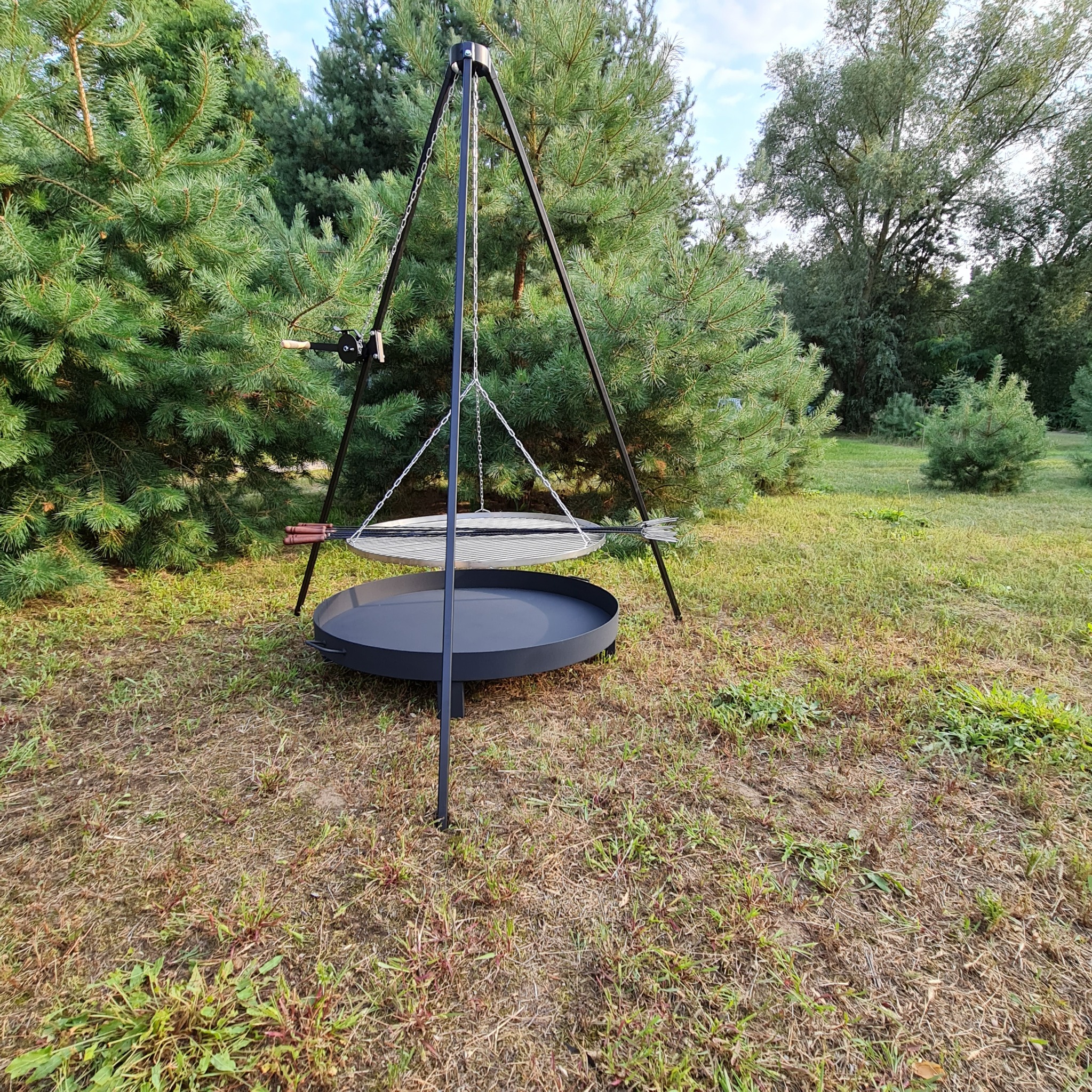 Metalowy grill trójnożny z rusztem i paleniskiem, ustawiony na trawniku przed drzewami iglastymi.