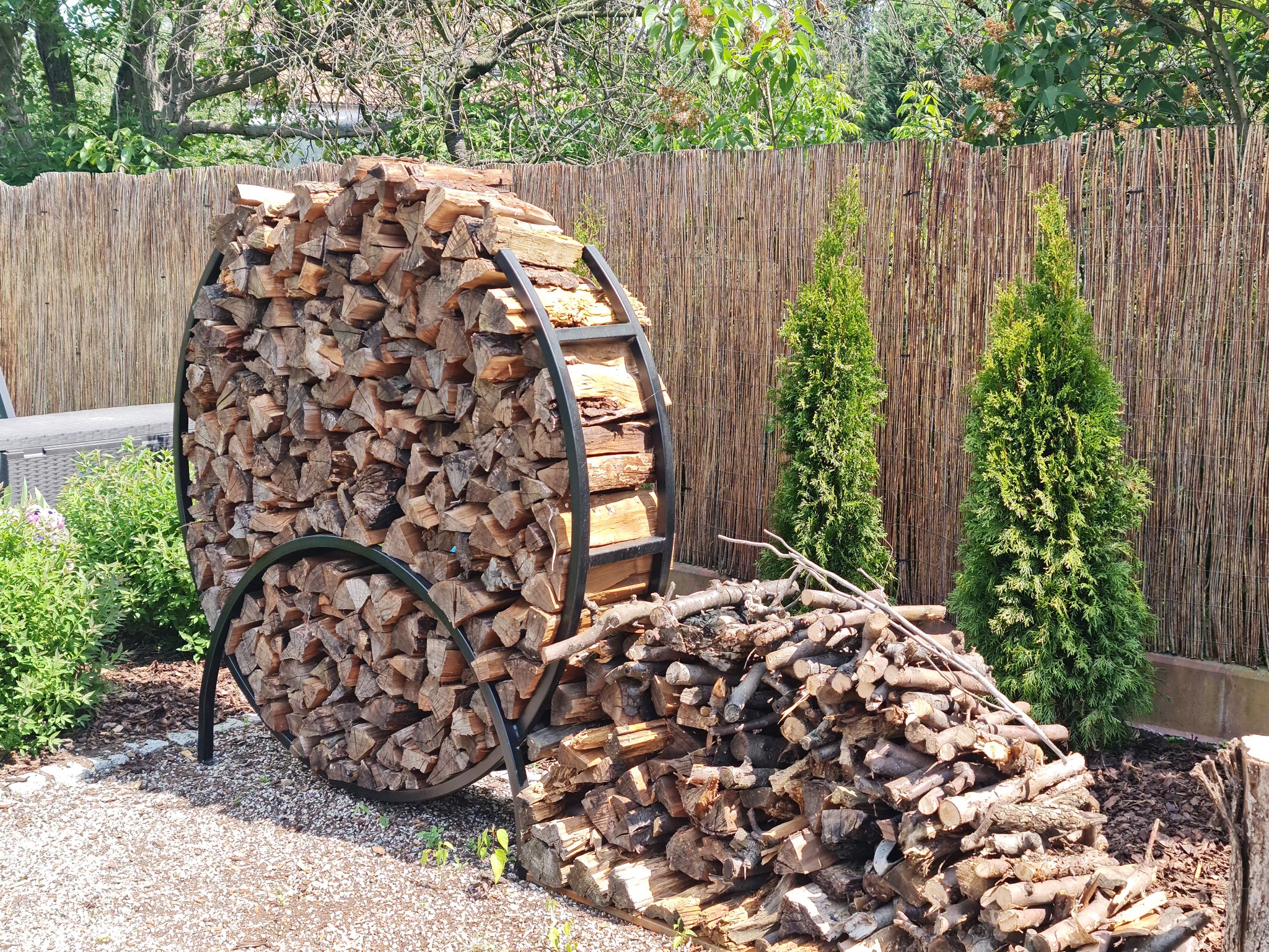 Okrągły, metalowy regał wypełniony pociętym drewnem opałowym, na tle plecionej maty osłonowej i dwóch małych tujek.