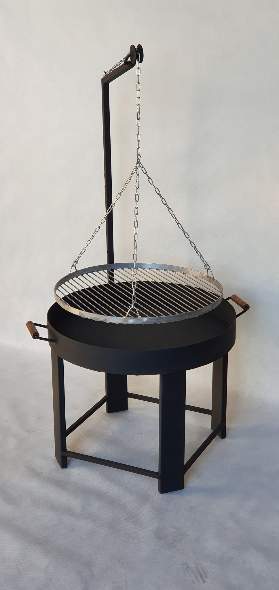 Czarny, metalowy grill z rusztem zawieszonym na łańcuchach, z drewnianymi uchwytami po bokach, stojący na czterech nogach.