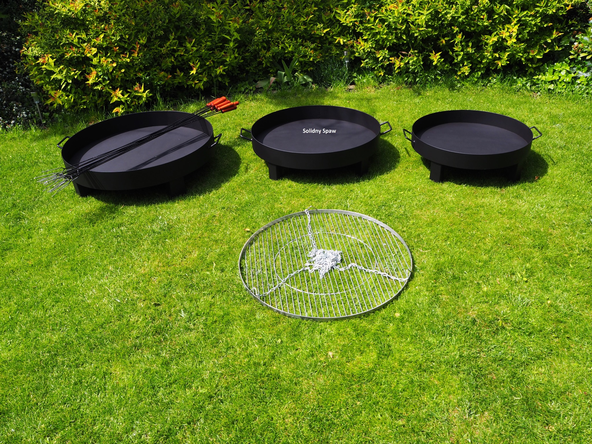 Trzy czarne, metalowe paleniska ogrodowe o różnych rozmiarach, z rusztem i szpikulcami do szaszłyków, rozłożone na trawie przed krzewem.