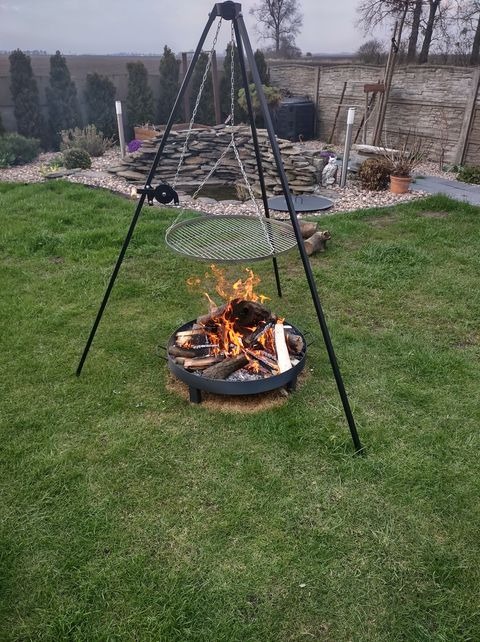 Metalowe palenisko ogrodowe na trójnogu z rusztem, płonące drewno w palenisku, w tle trawnik, kamienny ogródek i ogrodzenie.
