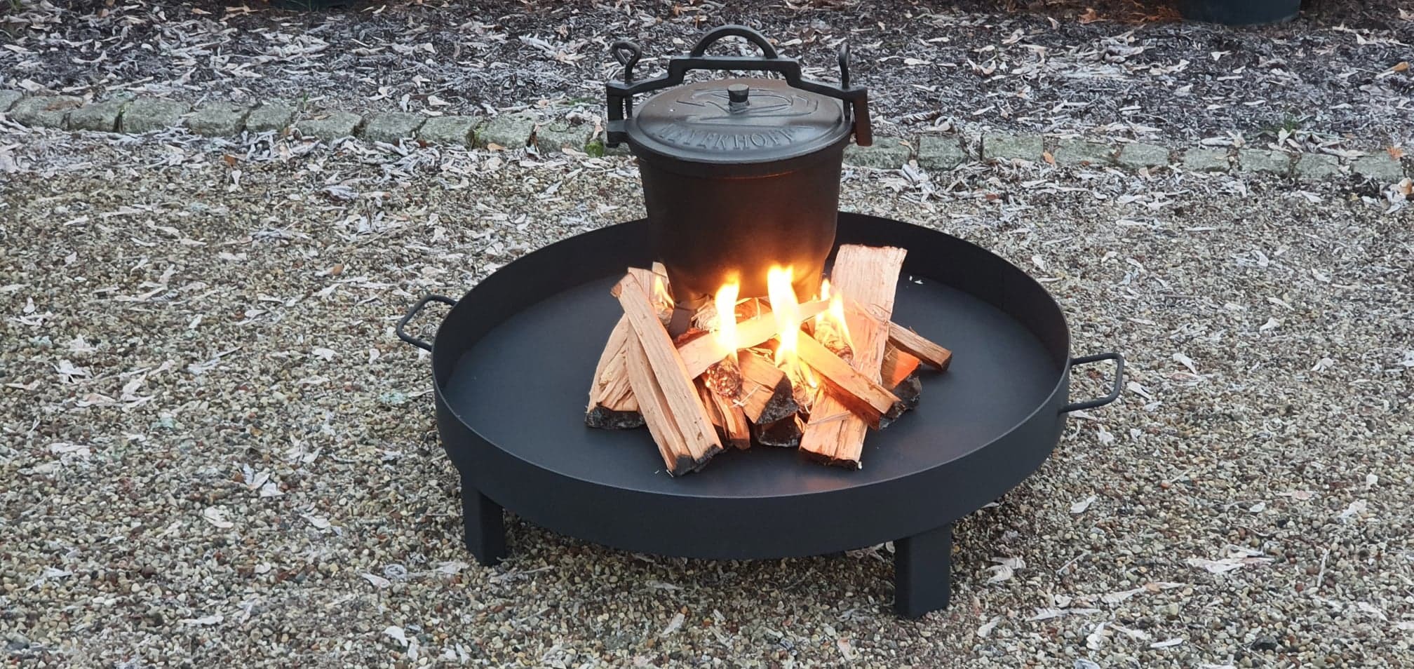 Czarny, metalowy palenisko ogrodowe z palącym się drewnem i wiszącym nad nim kociołkiem, ustawione na żwirowym podłożu.