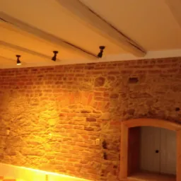 Wnętrze z fragmentem ściany wykończonej cegłą i kamieniem, oświetlone punktowymi lampami sufitowymi i wiszącą lampą przemysłową na łańcuchu, z widocznym drewnianym stropem i wnęką w ścianie.
