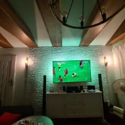 Pokój dzienny z telewizorem, kamienna ściana podświetlona na zielono, drewniane belki na suficie i lampa z liną, w tle zasłony.