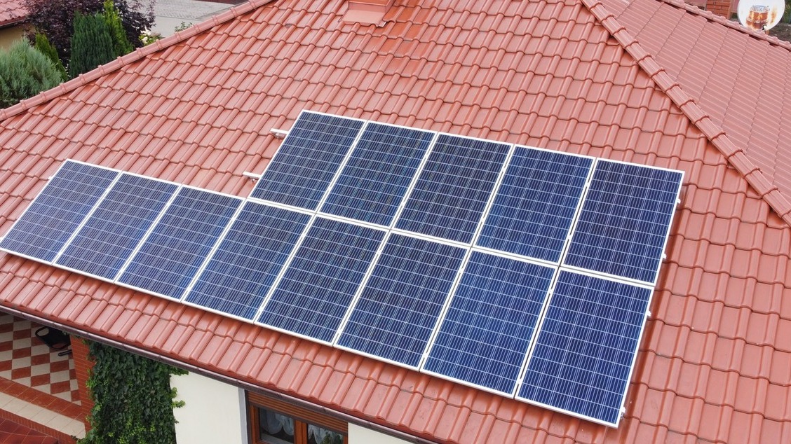 Panele słoneczne na dachu z czerwonej dachówki, widok z góry, biała elewacja budynku w tle.