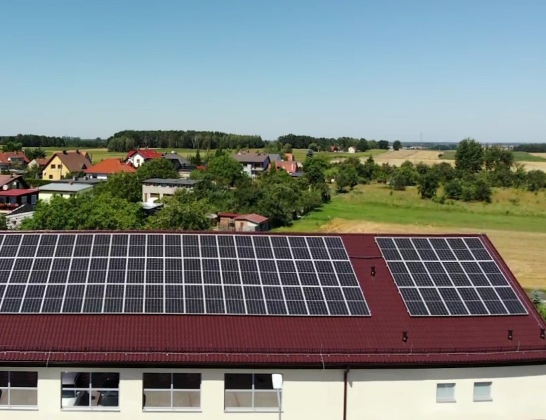 Panele słoneczne na dachu budynku z czerwonej blachy, widok z góry na tle krajobrazu wiejskiego.