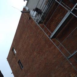 Daniel - Mur z czerwonej cegły w trakcie ocieplania, widoczne rusztowanie i pracownik na wysokości, fragmenty styropianu na górnej krawędzi ściany.