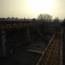 Zbrojenie fundamentów w trakcie budowy, widoczne szalunki, stalowe pręty, narzędzia i krzesła na placu budowy w pochmurny dzień.